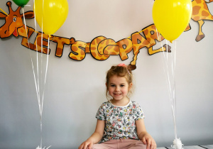 Dziewczynka siedzi na tle fotograficznym i pozuje do zdjęcia wśród balonów.